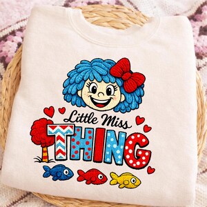 May include: A cream-coloured sweatshirt with a cartoon illustration of a girl with blue hair and a red bow. The text "Little Miss THING" is printed below the girl, along with red hearts and colourful fish.