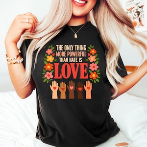 Può includere: T-shirt nera con la scritta "THE ONLY THING MORE POWERFUL THAN HATE IS LOVE" in bianco e rosso. Il design include una ghirlanda floreale e mani di varie tonalità di pelle con simboli di cuore.