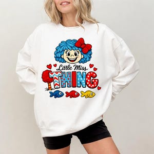 May include: White sweatshirt with a cartoon graphic of a character with blue hair, a red bow, and the text "Little Miss THING". The design includes red hearts and three colourful fish.