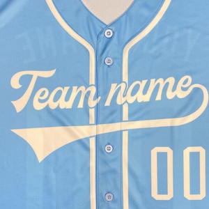 May include: A light blue baseball jersey featuring cream-colored piping along the button placket and neckline. The text "Team name" is printed in a cream script font across the chest, accompanied by the number "00" in a cream block font.