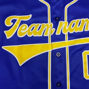 May include: A royal blue button-down baseball jersey featuring the text Team name in yellow script with white trim. The jersey includes a yellow swoosh graphic across the front, vertical yellow and white piping, and dark blue buttons.