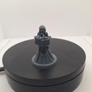 May include: A gray miniature figurine of a robed figure holding a basket. The figure has a hooded cloak and a long, flowing gown. The basket is intricately detailed. The figurine is on a black rotating platform.