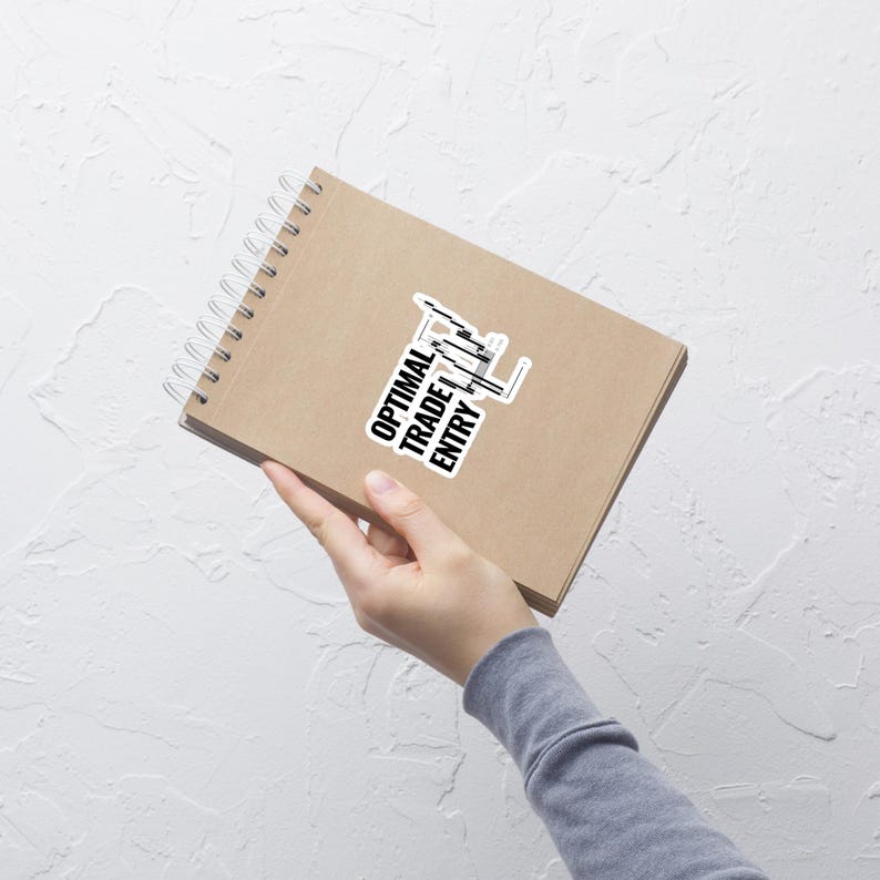 Pu&ograve; includere: Un quaderno marrone con un adesivo bianco che dice "OPTIMAL TRADE ENTRY" con un grafico a candele in bianco e nero.