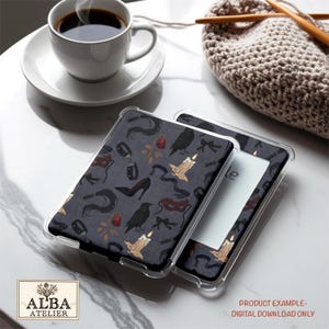 Puede incluir: Funda para Kindle con un fondo gris oscuro con un patr&oacute;n de cuervos negros, tacones altos, velas y otros elementos g&oacute;ticos. La funda se muestra junto a una taza de caf&eacute; y un objeto de ganchillo.