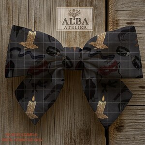 Puede incluir: Una pajarita grande, de color gris oscuro, con un patr&oacute;n repetido de velas, l&aacute;tigos y otros elementos g&oacute;ticos. La pajarita est&aacute; colocada sobre un fondo de madera y presenta el logotipo "ALBA ATELIER".