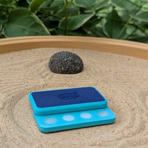 May include: A small zen garden with a dark gray stone and a blue and teal magnetic sand rake. The rake has a textured surface and three white circles. The sand is tan and has a pattern.