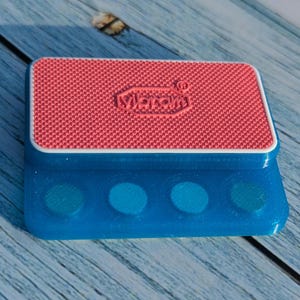 Op de afbeelding: Een blauw en rood rechthoekig object met het woord "Vibram" in reliëf op het rode oppervlak. Het object heeft vier blauwe cirkelvormige inkepingen aan de voorkant. Het object ligt op een houten oppervlak.