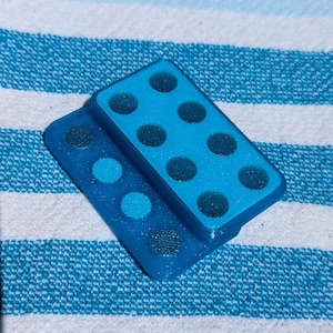 May include: Two blue rectangular game pieces with black dots, resembling dominoes, are stacked on a blue and white striped surface. The top piece has six dots, the bottom piece has two dots.