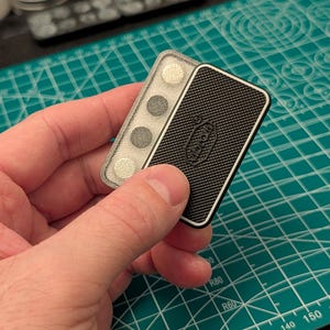 Puede incluir: Un objeto rectangular en blanco y negro con una superficie texturizada y un borde negro. El objeto tiene la palabra "Vibram". Junto a él, un objeto rectangular transparente con tres círculos grises.