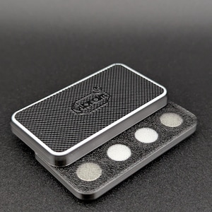 Puede incluir: Un estuche rectangular con un patrón de fibra de carbono en la tapa y bordes plateados. La tapa está ligeramente abierta, revelando una bandeja con cuatro objetos circulares metálicos. El estuche está sobre un fondo oscuro.