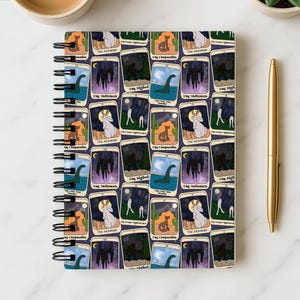 May include: Spiral-bound notebook featuring a repeating pattern of tarot card illustrations. The cards depict various cryptids like the Mothman and the Jackalope, set against a dark blue background. A gold pen and a cup of coffee are also visible.