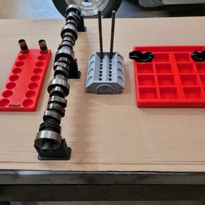 May include: An assortment of automotive engine parts, including a camshaft, valve guides, and red and gray storage trays. The camshaft is silver and black, and the trays are labeled "FRONT". The parts are arranged on a cardboard surface.