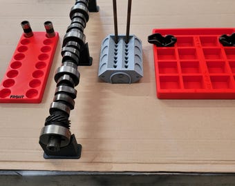May include: An assortment of automotive engine parts, including a camshaft, valve guides, and red and gray storage trays. The camshaft is silver and black, and the trays are labeled "FRONT". The parts are arranged on a cardboard surface.