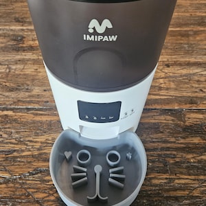 Peut inclure: Distributeur automatique de nourriture pour animaux de compagnie, noir et blanc, avec la marque "IMIPAW". Le bol est gris et en forme de visage de chat. Idéal pour contrôler les portions.