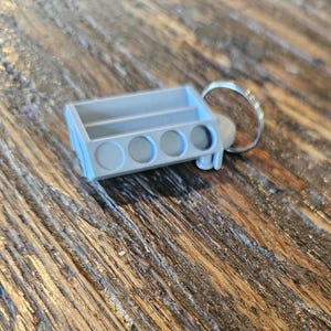 May include: A gray, 3D-printed keychain featuring a miniature engine block design. The keychain has four circular openings and a rectangular top, attached to a silver keyring. The item is resting on a wooden surface.
