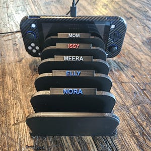May include: A black gaming console stand with slots labeled "MOM", "ISSY", "MEERA", "ELLY", and "NORA". A gaming console with a carbon fiber design is docked in the stand. The stand is on a wooden surface.