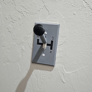 May include: A gray plastic gear shifter with a black knob and a silver lever. The shifter has the numbers 1, 2, 3, 4, and R printed on it.