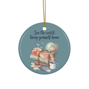 May include: A round, blue ceramic ornament hanging from a gold string. It features the text "See the world, bring yourself home" above a watercolor illustration of a brown suitcase, a globe, a passport, a coffee mug, and flying birds.
