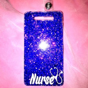 Puede incluir: Un porta-credenciales rectangular azul brillante con la palabra "Nurse" en escritura blanca y un gráfico de estetoscopio blanco. El porta-credenciales tiene una correa de plástico transparente y un cierre de metal.