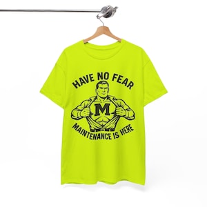 May include: A neon yellow t-shirt with a black graphic. The graphic features a superhero-like figure with the letter "M" on the chest, and the text "HAVE NO FEAR MAINTENANCE IS HERE". The shirt is hanging on a wooden hanger.