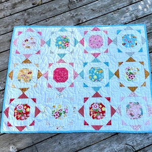 May include: A white quilt with a blue border. The quilt is made up of 12 squares, each with a different floral pattern in shades of pink, red, yellow, and blue. The squares are arranged in a grid pattern.