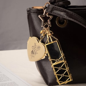 Può includere: Un portachiavi in metallo dorato attaccato a una borsa nera. Il portachiavi presenta un ciondolo a forma di stella, un medaglione a cuore inciso con un motivo floreale e il nome Sophia, e un porta rossetto con una cornice a griglia dorata.