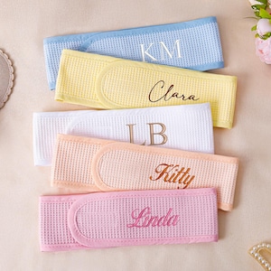 Op de afbeelding: Vijf gepersonaliseerde spa-hoofdbanden in pastelkleuren: lichtblauw, geel, wit, perzik en roze. Elke hoofdband heeft geborduurde tekst: "KM", "Clara", "LB", "Kitty" en "Linda". De hoofdbanden zijn gemaakt van wafelstof.
