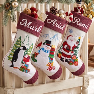 Op de afbeelding: Drie crèmekleurige kerstsokken met bordeauxrode, getextureerde boorden met de namen Mark, Ariel en Remi. De sokken zijn versierd met geborduurde scènes van een pinguïn, een sneeuwpop en de Kerstman met cadeautjes.