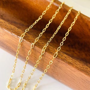 May include: Four delicate gold-coloured chain necklaces with rectangular links are arranged in a wooden tray. The chains are displayed on a white surface, with a small gold sequined object in the background.