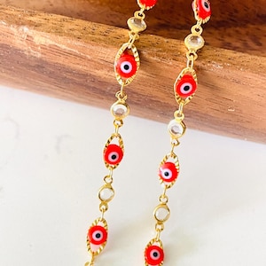 Könnte beinhalten: Zwei goldfarbene Ketten mit roten und weißen Evil-Eye-Anhängern und klaren Kristallakzenten. Die Ketten werden auf einer Holzoberfläche und einem weißen Hintergrund präsentiert. Die Evil-Eye-Anhänger sind rot mit einer schwarzen Pupille.