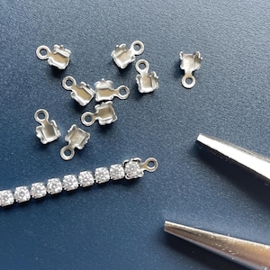 May include: A collection of small, silver-colored jewelry findings, including several open-backed settings and a section of a rhinestone chain. A pair of silver-colored pliers is also visible. The items are arranged on a dark blue surface.