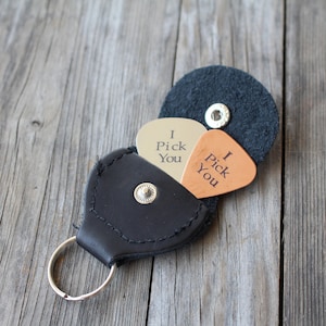 May include: A black leather keychain with a guitar pick holder. The holder has two guitar picks inside, one silver and one brown. The silver pick says "I Pick You" and the brown pick says "I Pick You".