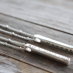 May include: Two silver bar necklaces with delicate chains. One bar has the name "Hannah" engraved on it, and the other has Roman numerals "XII V MMXV" engraved on it.