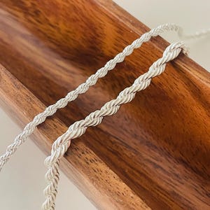 May include: Two silver rope chains of varying thicknesses are shown against a wooden background. The chains are made of twisted metal strands, creating a braided appearance. The chains are likely jewellery, such as necklaces or bracelets.