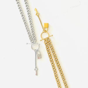 Może przedstawiać: Dwa naszyjniki z łańcuchami typu curb. Jeden jest w kolorze srebrnym z kłódką i zawieszką w kształcie klucza. Drugi jest w kolorze złotym z zawieszką w kształcie kłódki. Oba naszyjniki mają zapięcie typu T-bar.