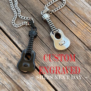 May include: Two guitar-shaped pendant necklaces. One is black, the other is silver. Both hang from a silver chain. The pendants are detailed with strings and sound holes. The text "CUSTOM ENGRAVED" is in red.
