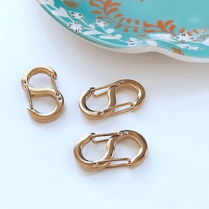 Peut inclure: Trois mousquetons dorés en forme de S sont présentés sur une surface blanche. Une assiette décorative turquoise avec un motif d'oiseau et floral est en arrière-plan. Les clips sont probablement pour des porte-clés ou des bijoux.