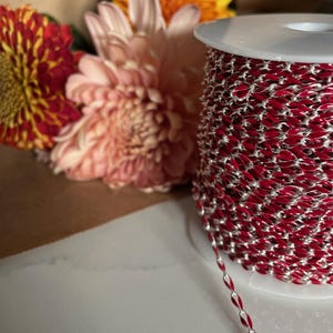 May include: A spool of red and silver chain with a white plastic spool. The chain is a flat, oval shape.