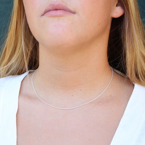 Puede incluir: Un collar de cadena de plata que se lleva alrededor del cuello de una persona. La cadena es fina y delicada.