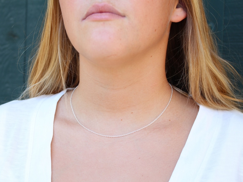 Puede incluir: Un collar de cadena de plata que se lleva alrededor del cuello de una persona. La cadena es fina y delicada.