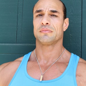 May include: A man with short dark hair is wearing a light blue tank top and a silver chain necklace with a small rectangular pendant. He is standing in front of a dark green wooden wall.