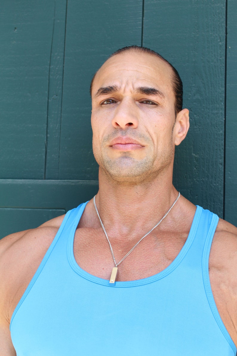 May include: A man with short dark hair is wearing a light blue tank top and a silver chain necklace with a small rectangular pendant. He is standing in front of a dark green wooden wall.
