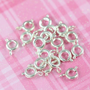 May include: A pile of silver-toned jump rings, also known as split rings, on a pink glittery background. These rings are used for attaching charms, pendants, and other jewelry components.