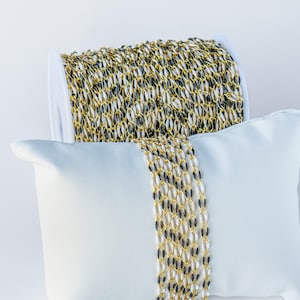 May include: A spool of gold and black chain, displayed on a white pillow. The chain is made of small, oval-shaped links.
