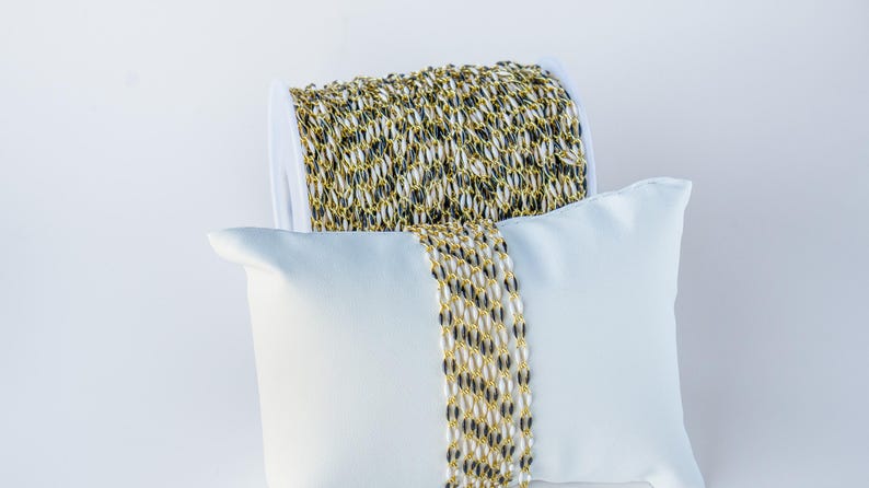 May include: A spool of gold and black chain, displayed on a white pillow. The chain is made of small, oval-shaped links.