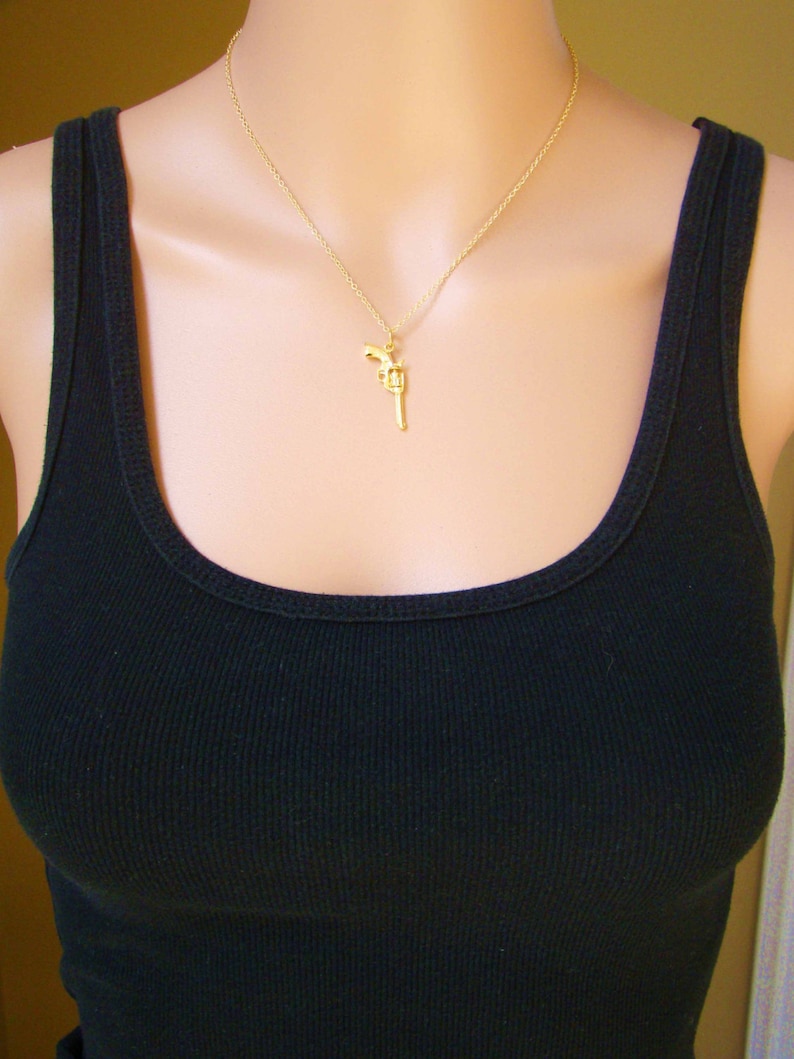Pode incluir: Um colar com pingente dourado com um pequeno pingente de rev&oacute;lver. O pingente pende de uma delicada corrente dourada. A pessoa est&aacute; usando uma regata preta canelada.