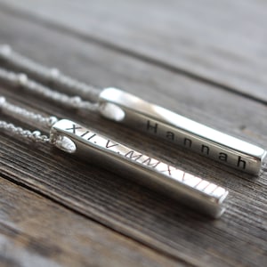 May include: Two silver bar necklaces with a heart-shaped cutout. One necklace has the Roman numeral date "XII.V.MMXXI" engraved on the bar. The other necklace has the name "Hannah" engraved on the bar.