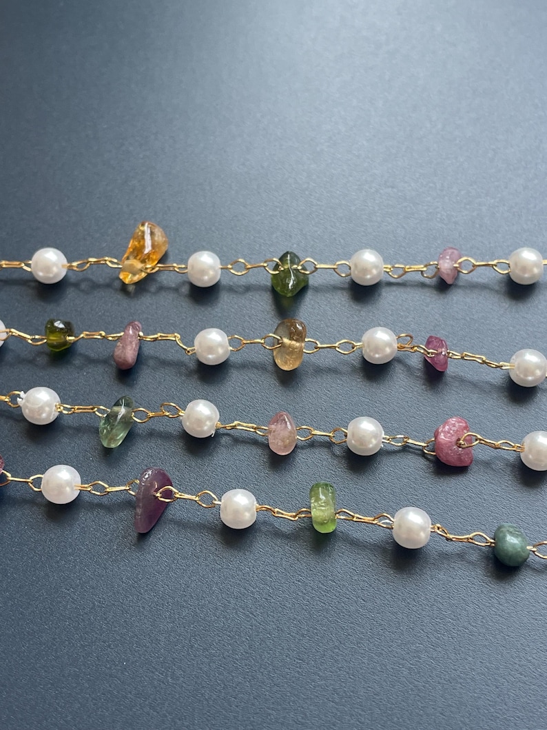 Puede incluir: Primer plano de varias hebras de joyer&iacute;a delicada. Cada hebra presenta una cadena dorada intercalada con perlas blancas y piedras preciosas de colores en tonos amarillos, verdes y rosas. El fondo es gris oscuro.