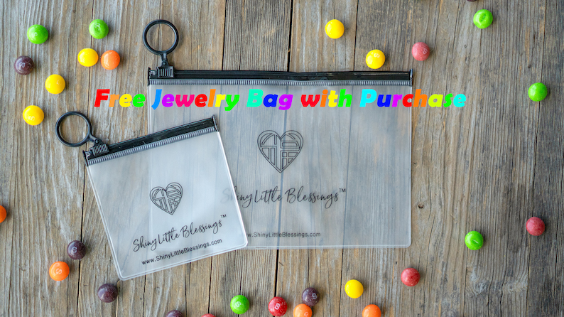 May include: Two clear plastic zippered bags with a black zipper and a black ring pull. The bags are labeled "Shiny Little Blessings" and feature a heart design. The text "Free Jewelry Bag with Purchase" is written in rainbow letters across the top of the image.