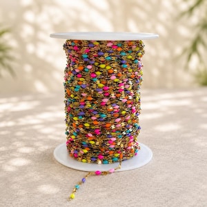 May include: A spool of gold-toned metal chain featuring small, oval-shaped beads in vibrant neon colors including pink, orange, yellow, blue, and purple. The chain is wrapped tightly around a white plastic spool, with a single strand extending downward.